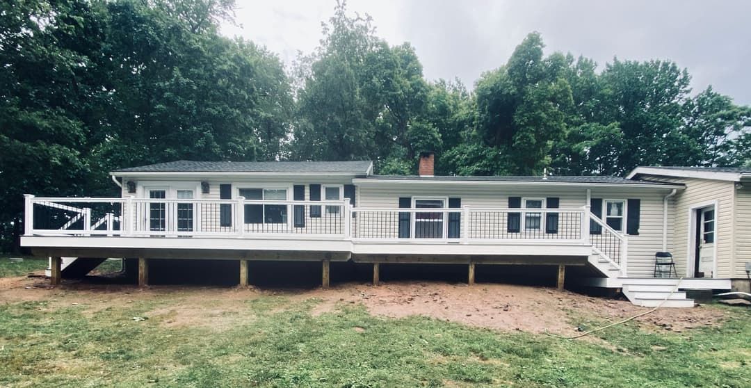 Ranch-style house with large deck, white siding, and windows. Green trees and grass in the background.