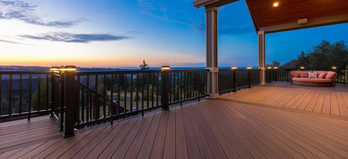Wooden deck overlooking a landscape at dusk. Lit railings surround the deck, and a couch sits under a covered area.