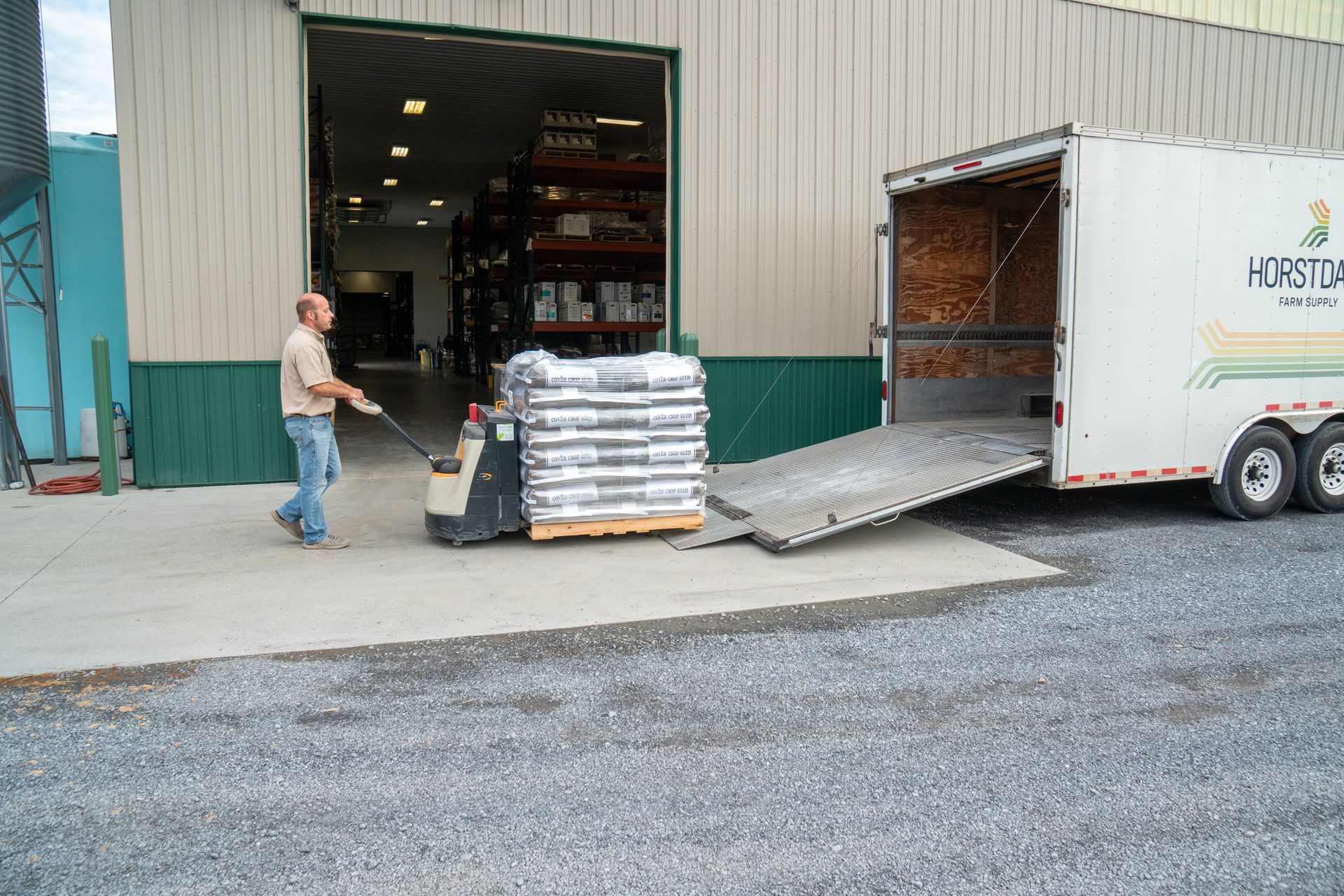 Man using pallet jack to load a pallet of bagged goods into a trailer at a warehouse.