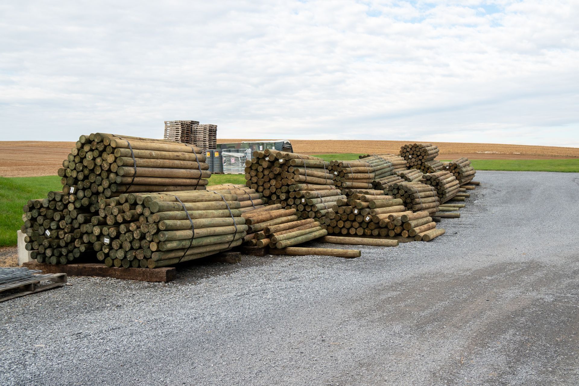 Piles of wooden fence posts on gravel, in front of a field and under a cloudy sky.