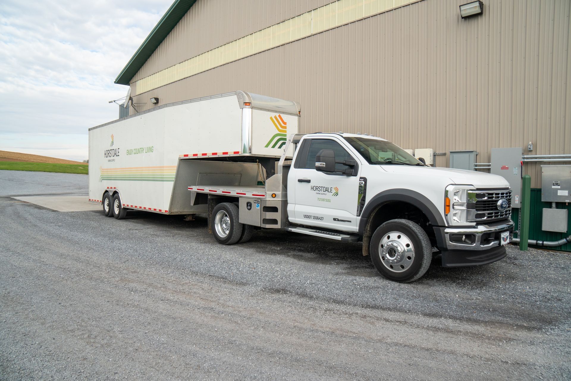 White truck with flatbed pulling a white trailer parked outside a beige industrial building on gravel.