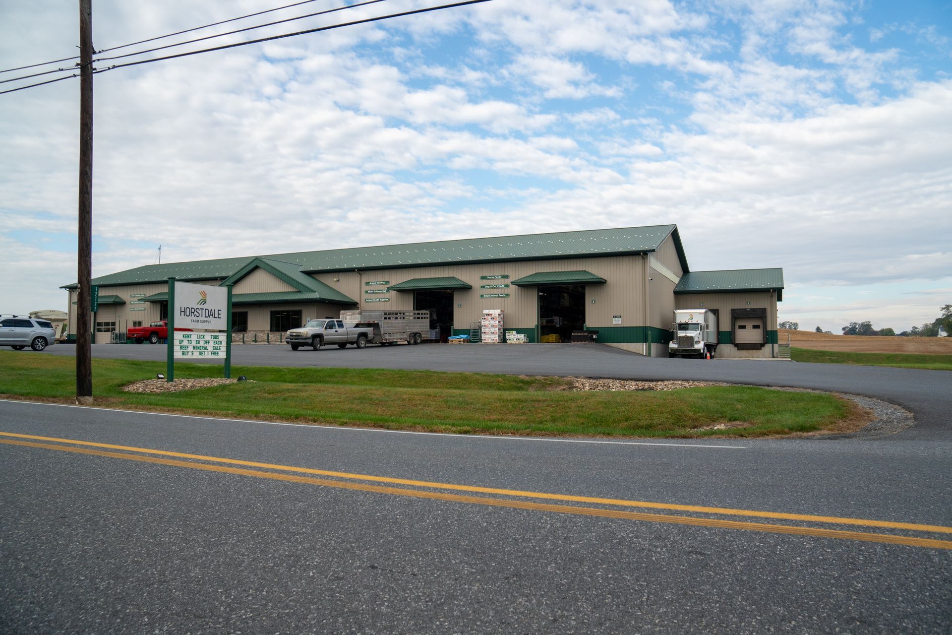 Large beige building with green trim, sign, and vehicles parked outside on a cloudy day.