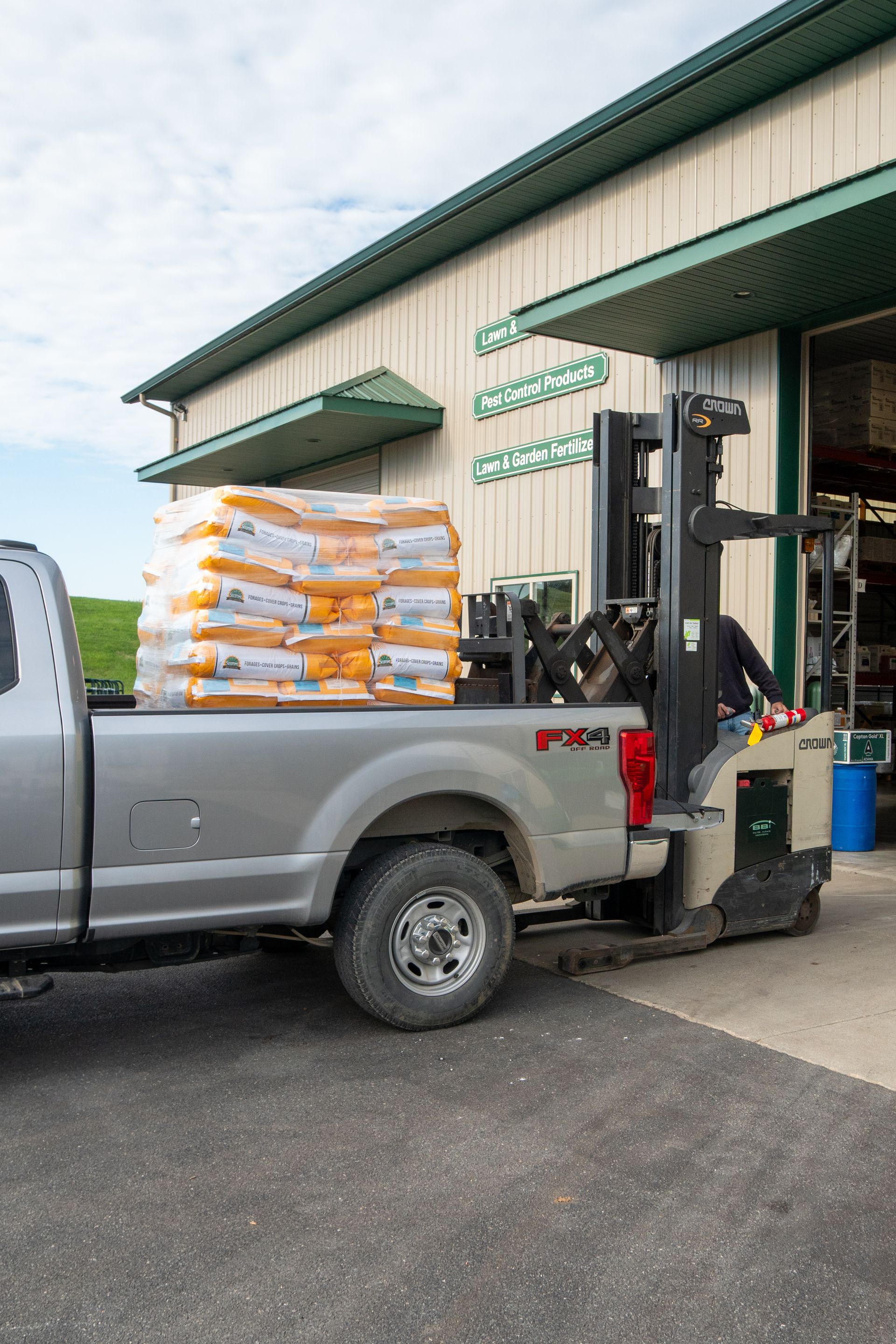 A forklift loading bags of product onto the back of a silver truck outside of a building.