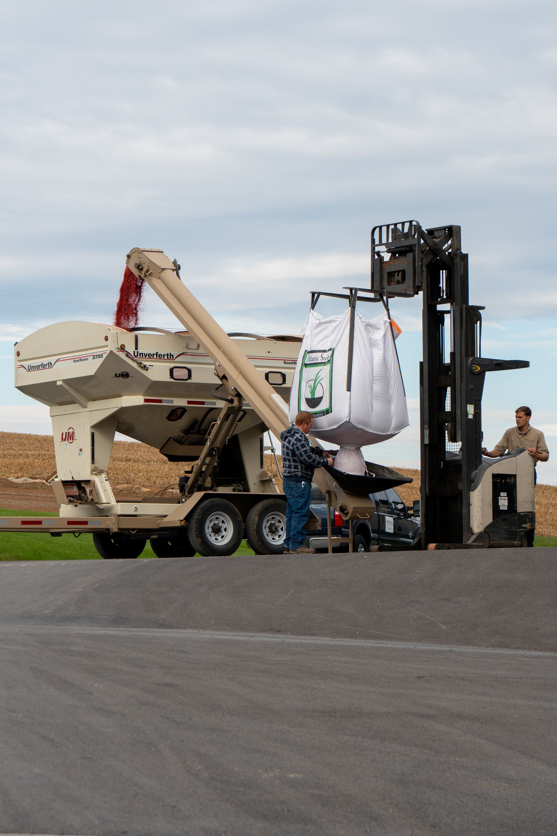 Workers loading a large bag with a forklift at a farm. Cream-colored trailer and overcast sky in the background.