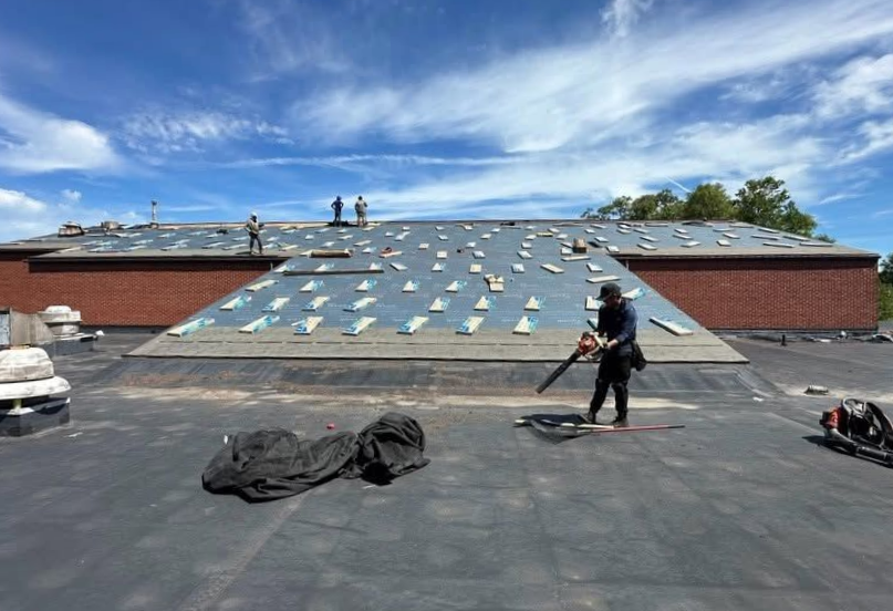 Roofer standing on a flat roof with underlayment, near red brick walls and a blue sky.