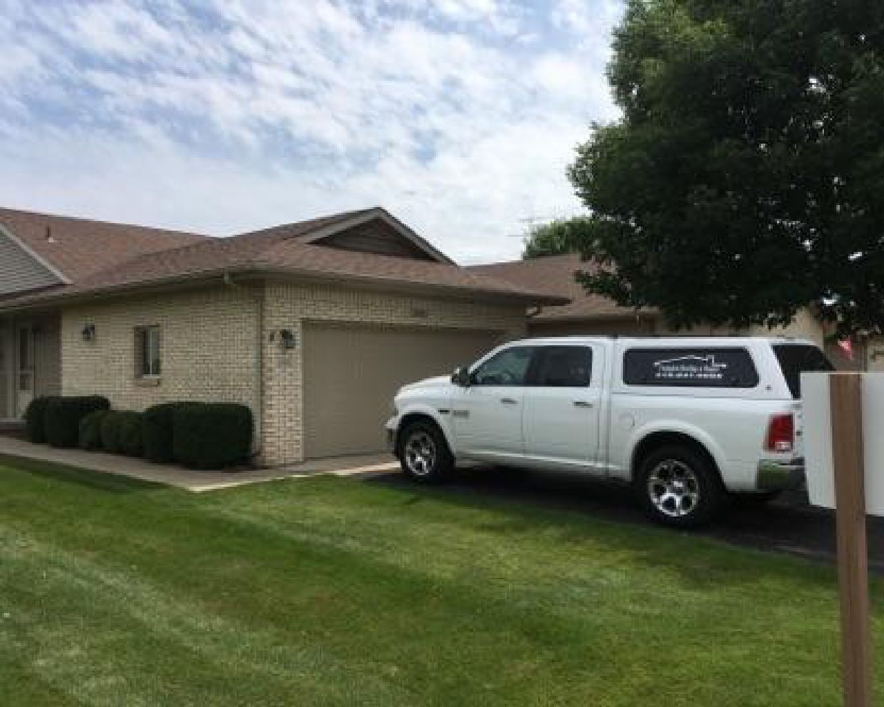 White pickup truck parked in front of a tan brick house with a garage and green lawn under a cloudy sky.