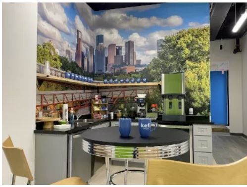 Office break room with skyline mural, coffee station, table, chairs, and blue door.