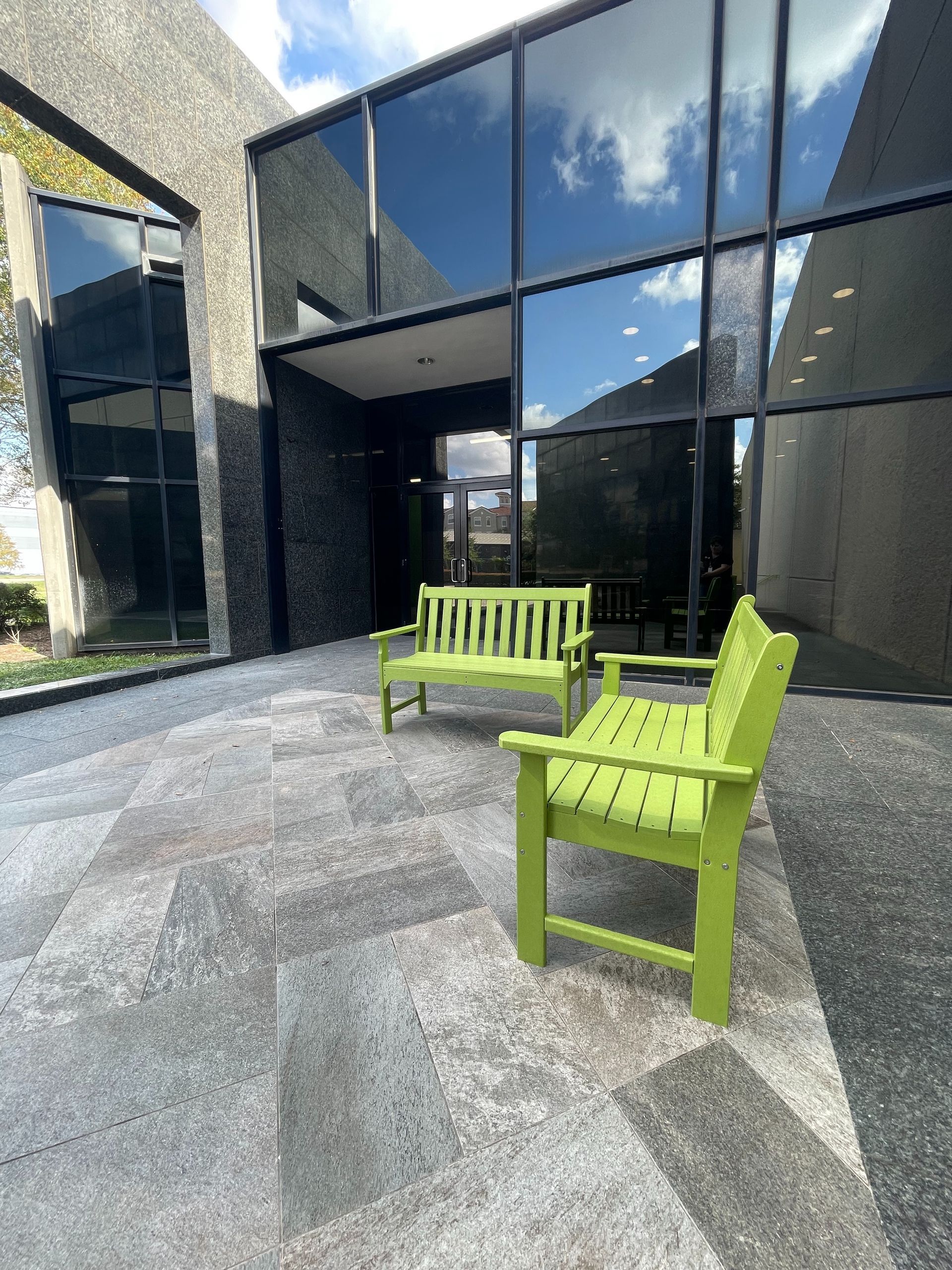 Two lime green benches sit on a gray stone patio in front of a modern building with large windows.