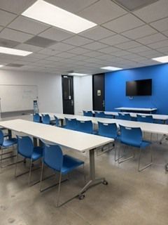 Empty classroom with white tables, blue chairs, tiled ceiling, and a wall-mounted TV on a blue accent wall
