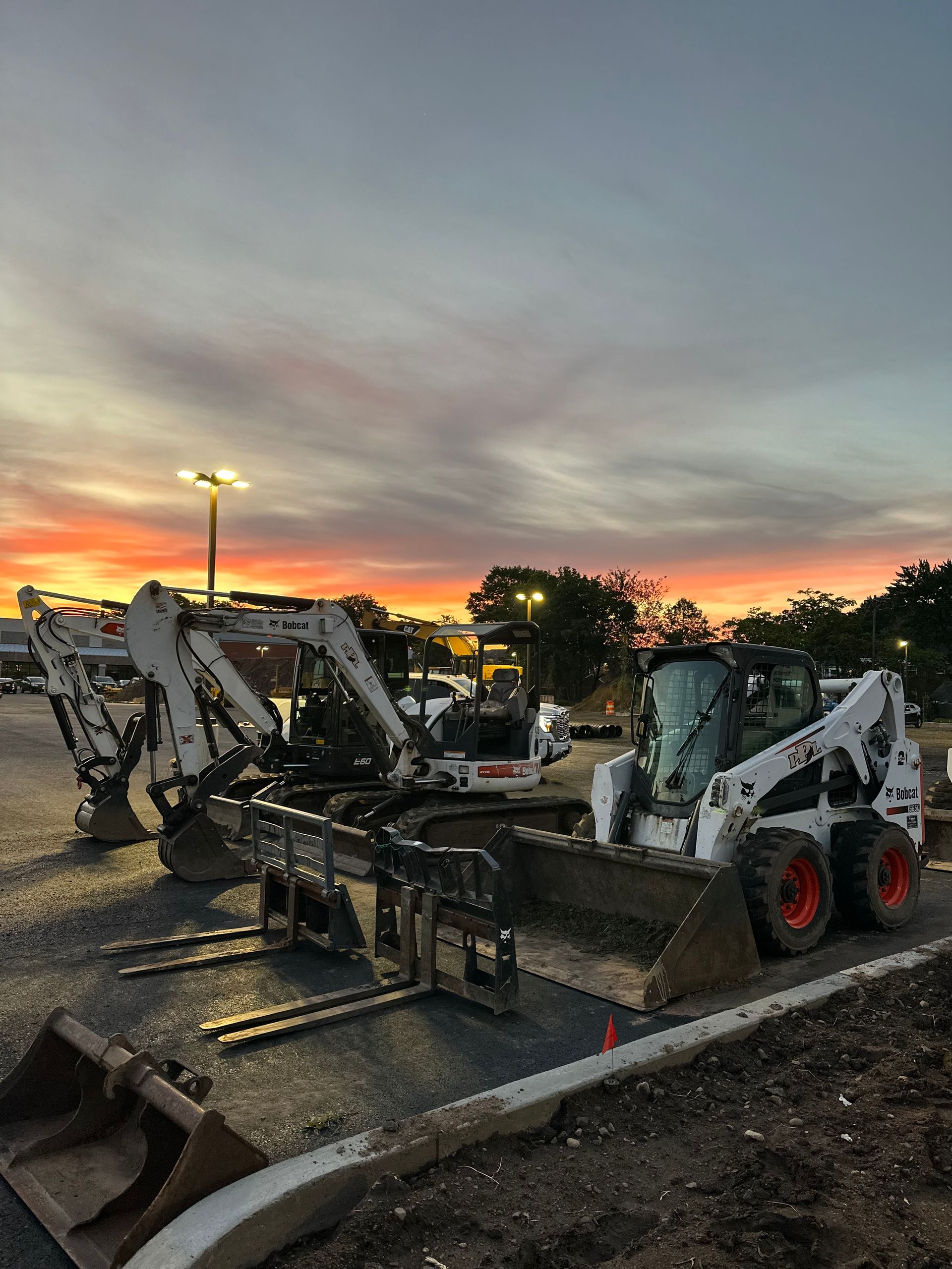 A row of construction vehicles are parked in a parking lot at sunset.