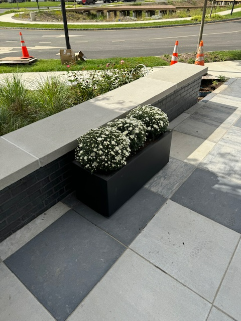 A black planter filled with white flowers sits on a tiled sidewalk.