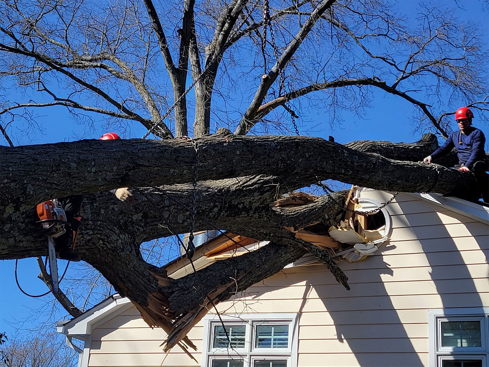 Workers in red helmets remove a fallen tree trunk from a house roof under a blue sky