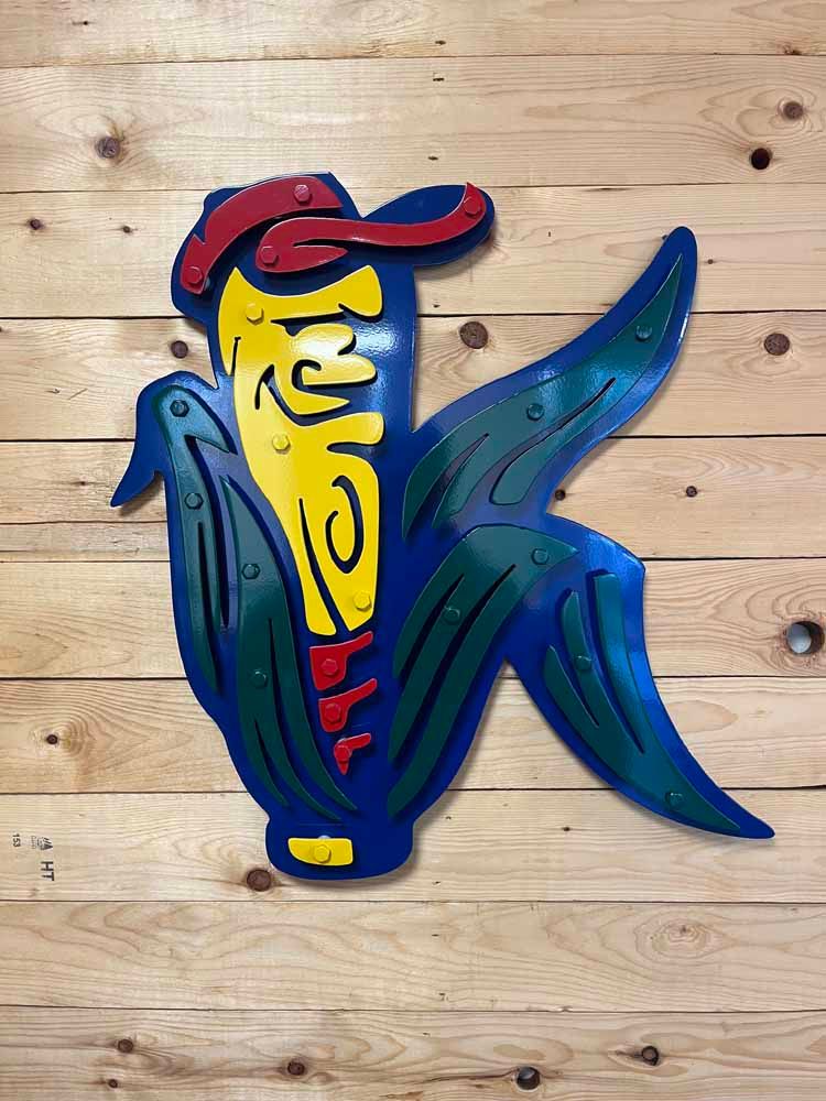 A colorful sign that looks like a corn on the cob is hanging on a wooden wall.