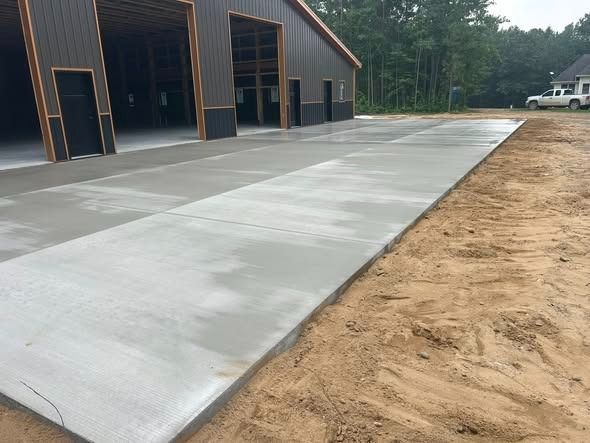 Newly poured concrete pad in front of a metal building with open bays, surrounded by dirt and a forested area.