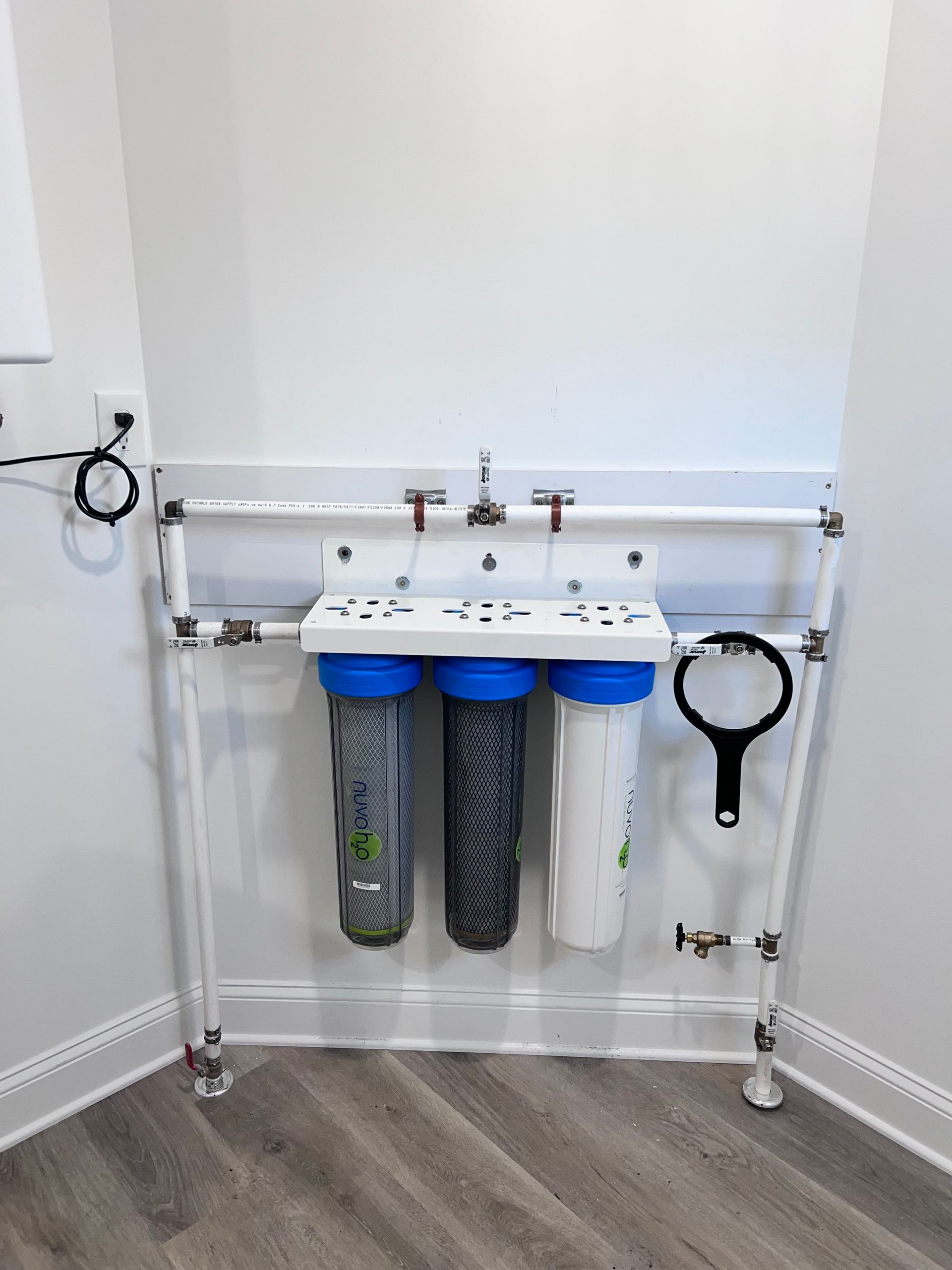 Water filter system mounted on a white pipe frame in a corner against a white wall.