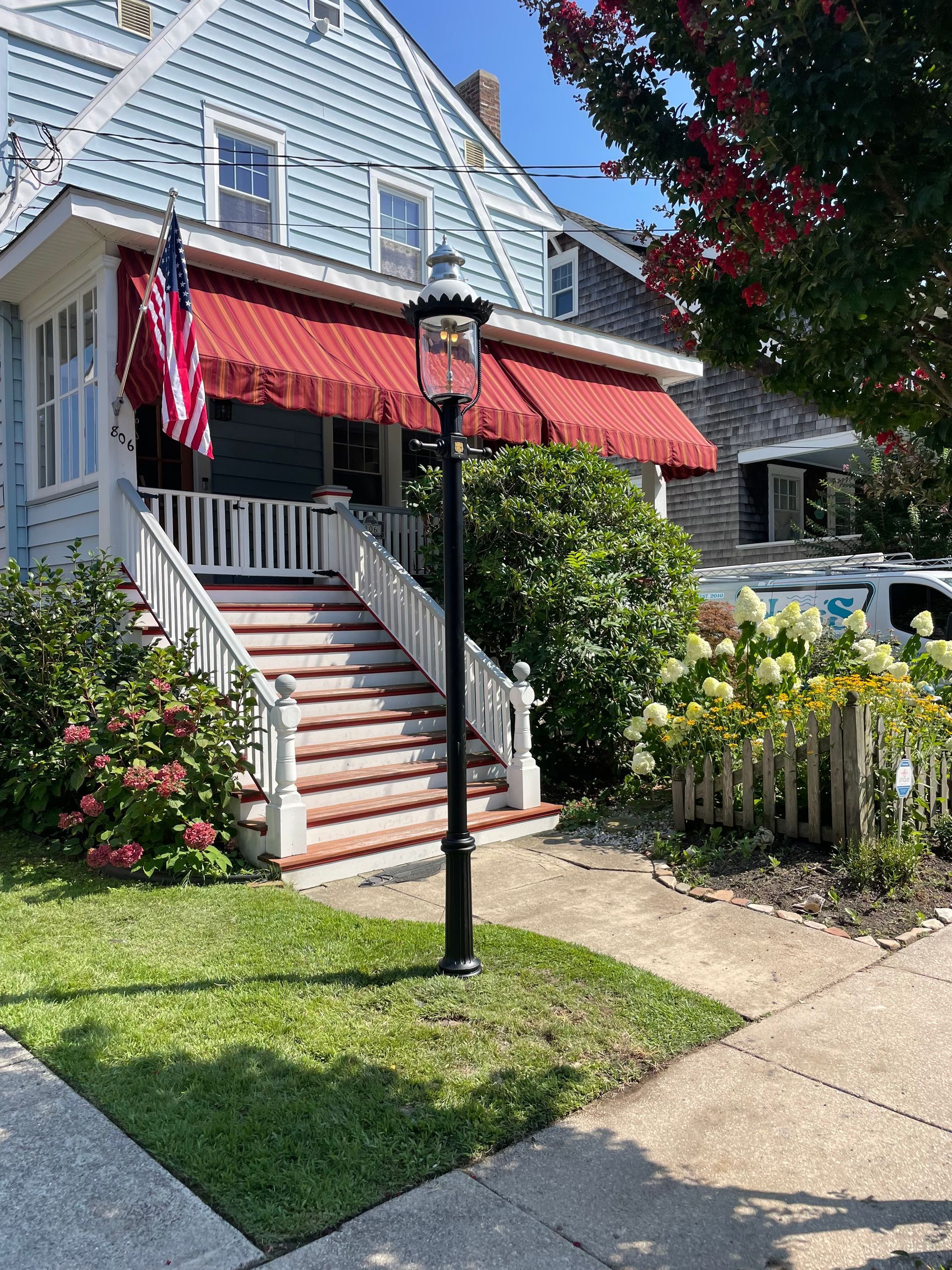 Blue house with red awning, American flag, white stairs, black lamp post, green lawn.