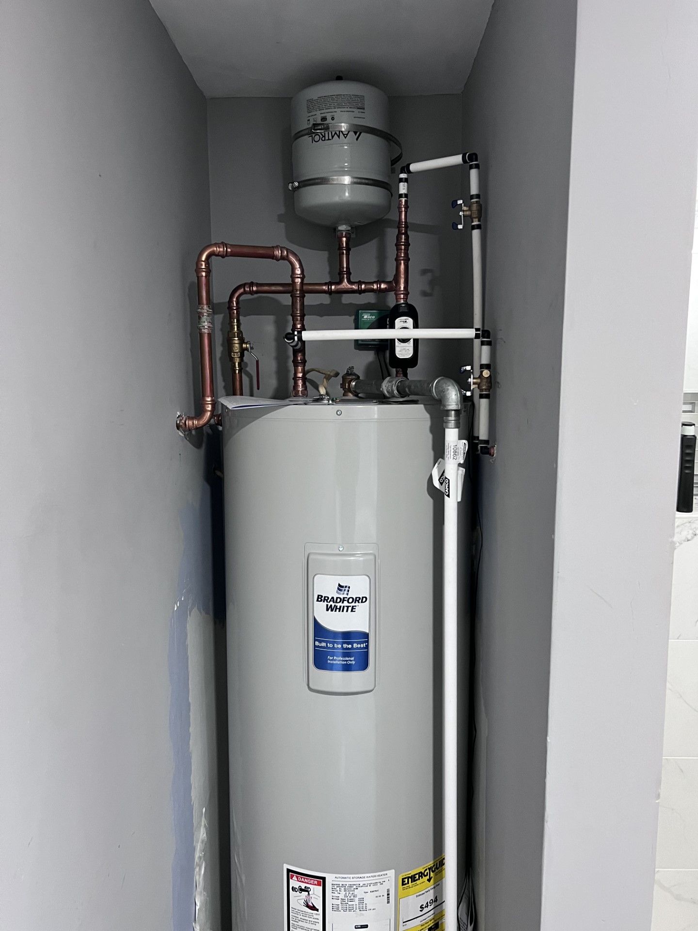 A tall, silver water heater with copper pipes and an expansion tank in a small alcove.