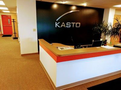KASTO office reception area with the company logo on a black wall, front desk with orange and white accents, and a hallway to the left.
