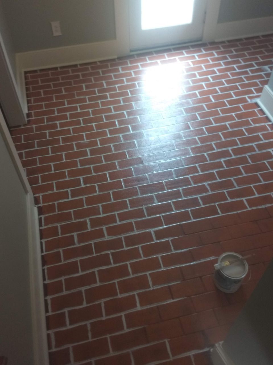 Brick pattern floor, reddish-brown bricks with white grout, leading to a door with sunlight.