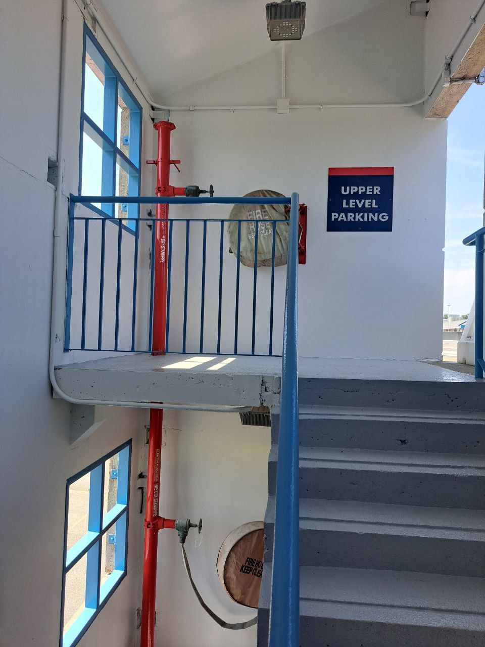 Staircase leading up with blue railing, red fire hose, white walls, and sign reading 