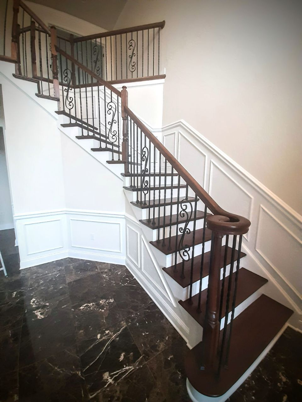Elegant two-story staircase with dark wood steps and railing, black wrought iron balusters, and white paneled walls.