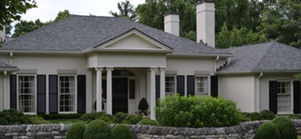 A white house with a dark roof and black shutters. Stone wall and green bushes in front.