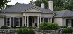 Beige-colored house with black shutters, columns, and a stone wall in front. Green trees and a gray roof.