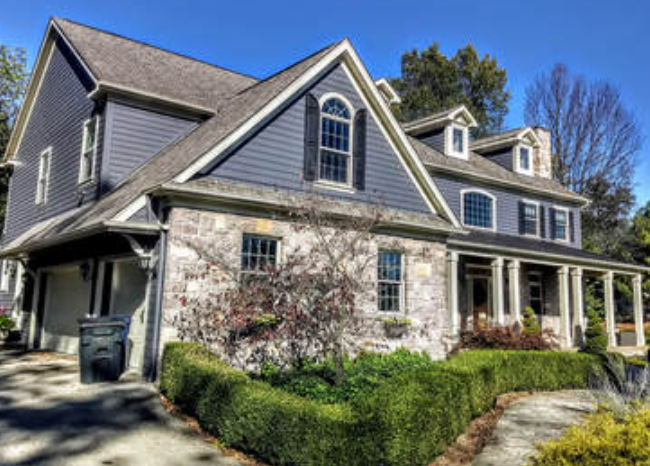 Two-story house with gray siding and stone exterior, front porch, and manicured landscaping under a blue sky.