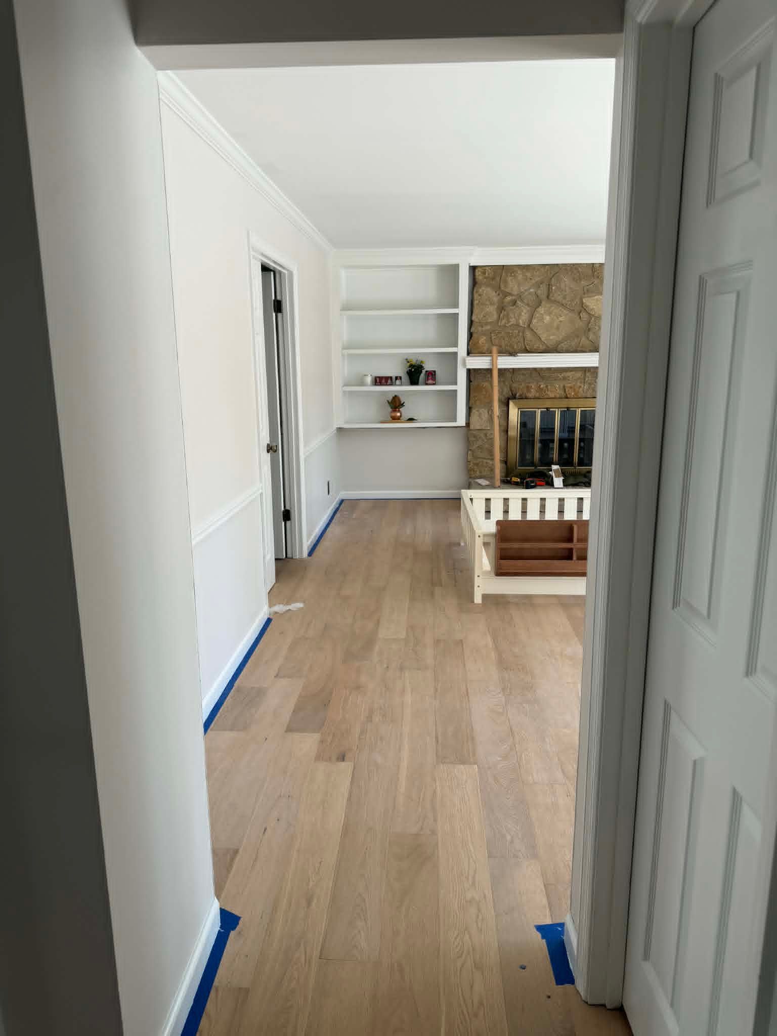Empty hallway opening into a room with light wood floors, white walls, and built-in shelves by a fireplace.
