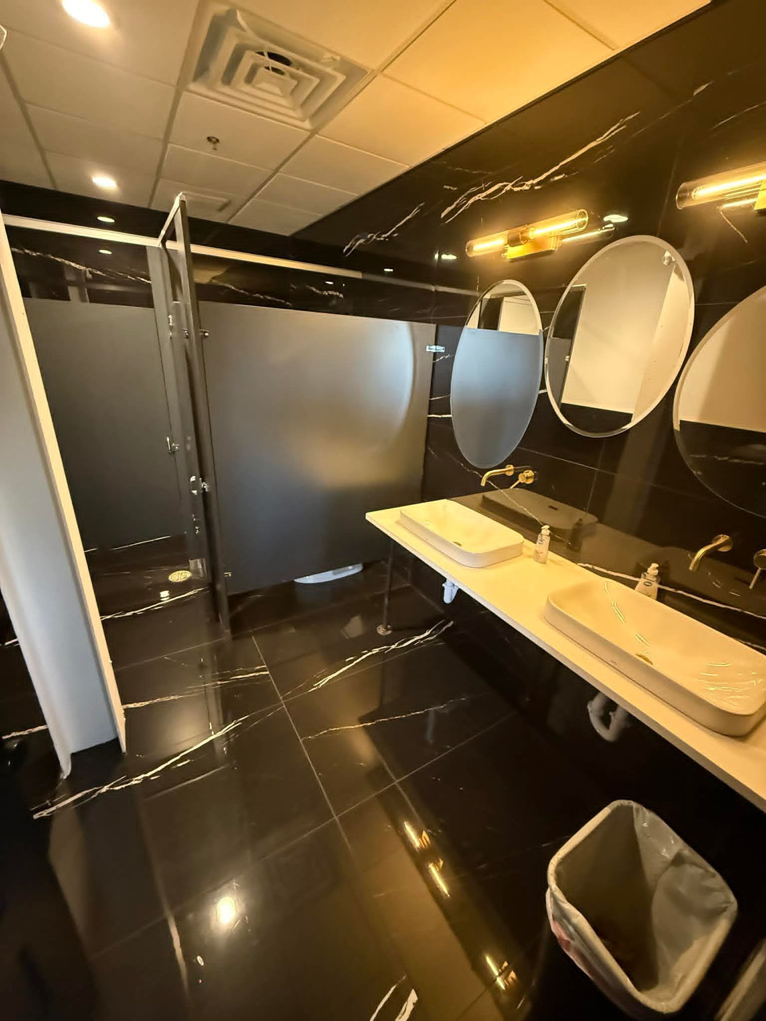 Modern restroom with black tile, sinks, mirrors, and partitioned stalls under warm lighting