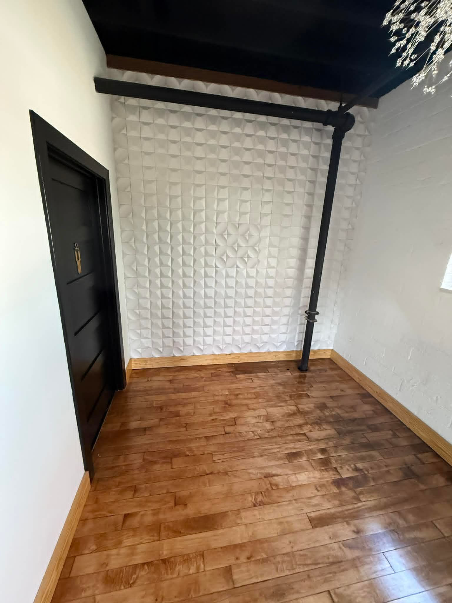 Small empty room with wood floor, black door, white textured wall, and dark ceiling beams.