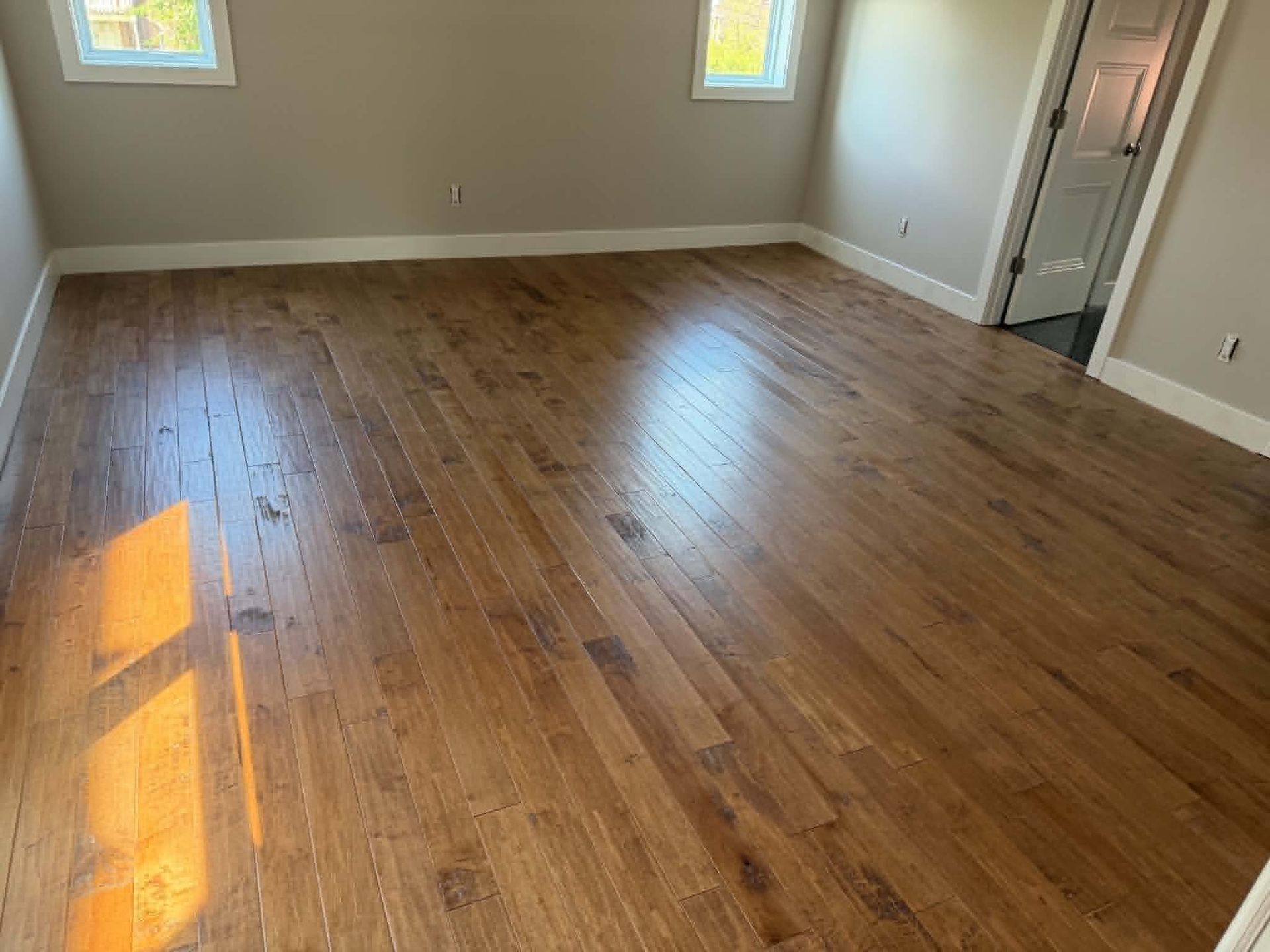 Empty room with hardwood floors, beige walls, and sunlight from two windows