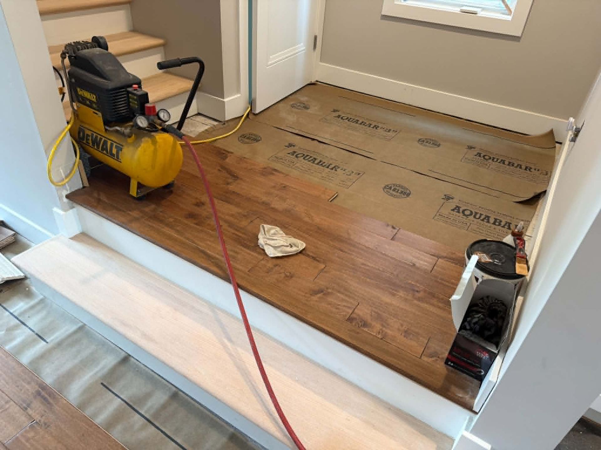 Partially renovated room with exposed subfloor, hardwood flooring, and a compressor with hose on the floor