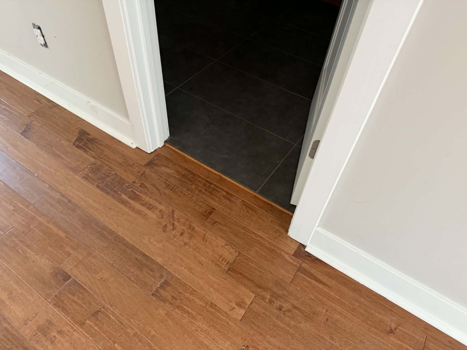Wood flooring meets a dark tiled doorway beside white walls and trim.