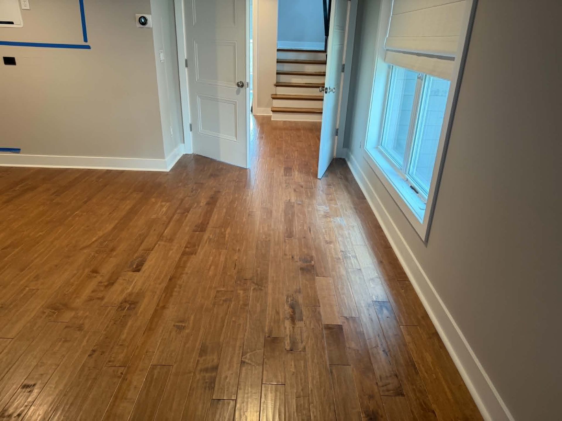 Empty hardwood-floored room with white walls, a doorway to stairs, and a large window on the right.