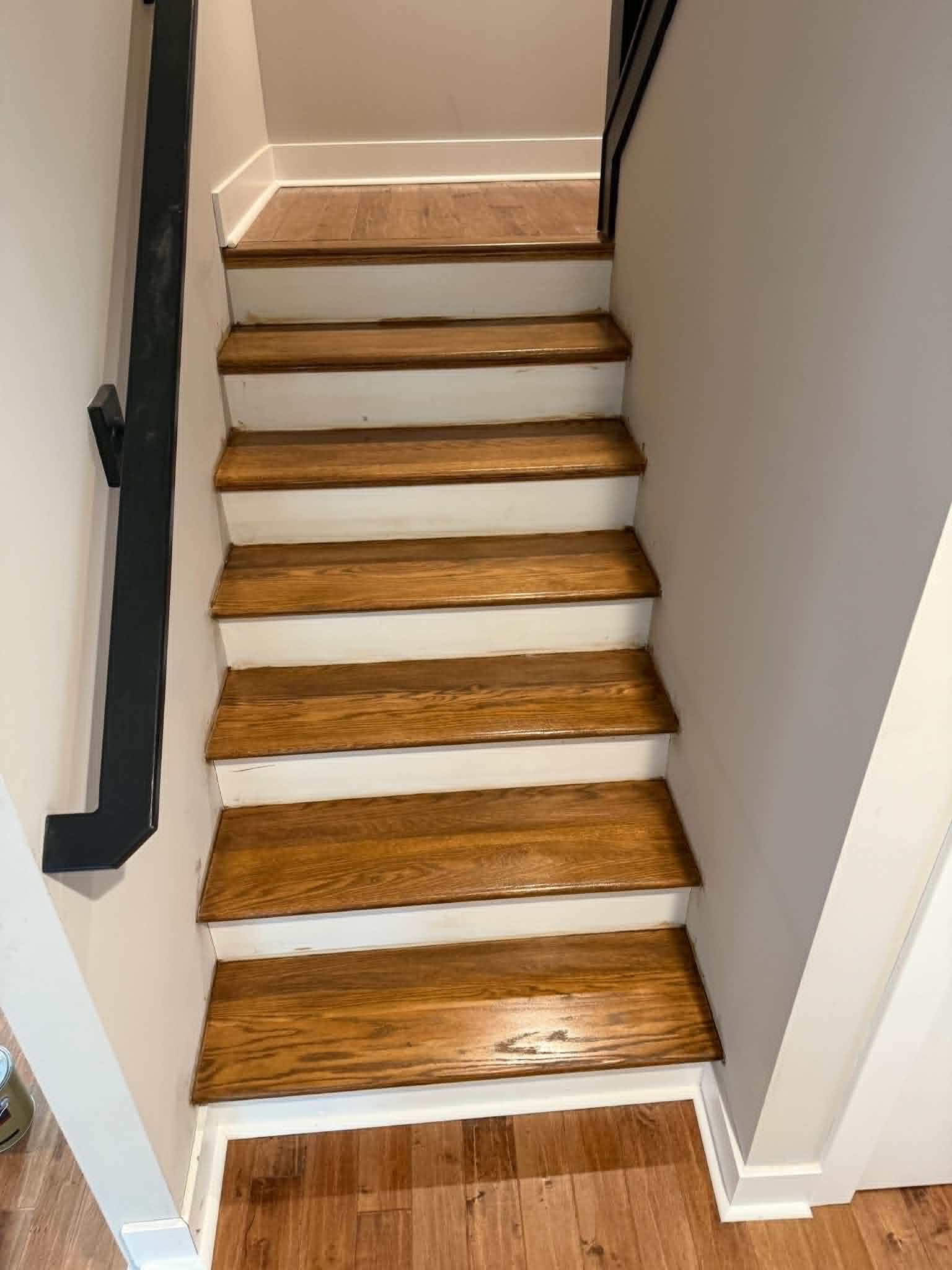Narrow indoor staircase with wooden steps, white risers, and black handrails against light walls.