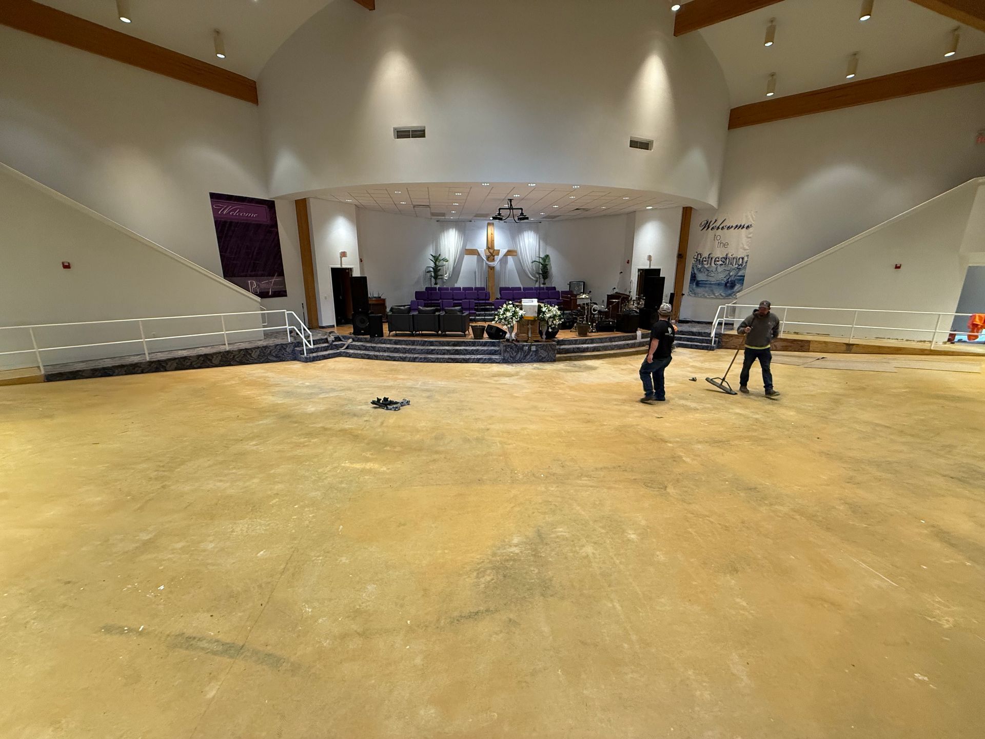 Two workers clean a large, bare, light-brown floor in a sanctuary with a raised stage featuring a cross and seating.