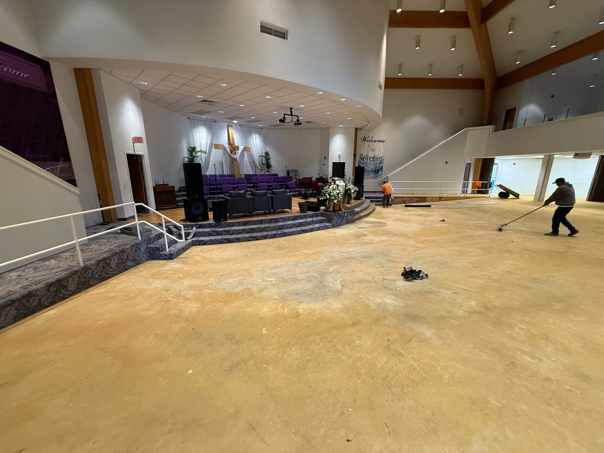 A worker cleans the bare concrete floor of a large sanctuary as another person stands near the raised stage.