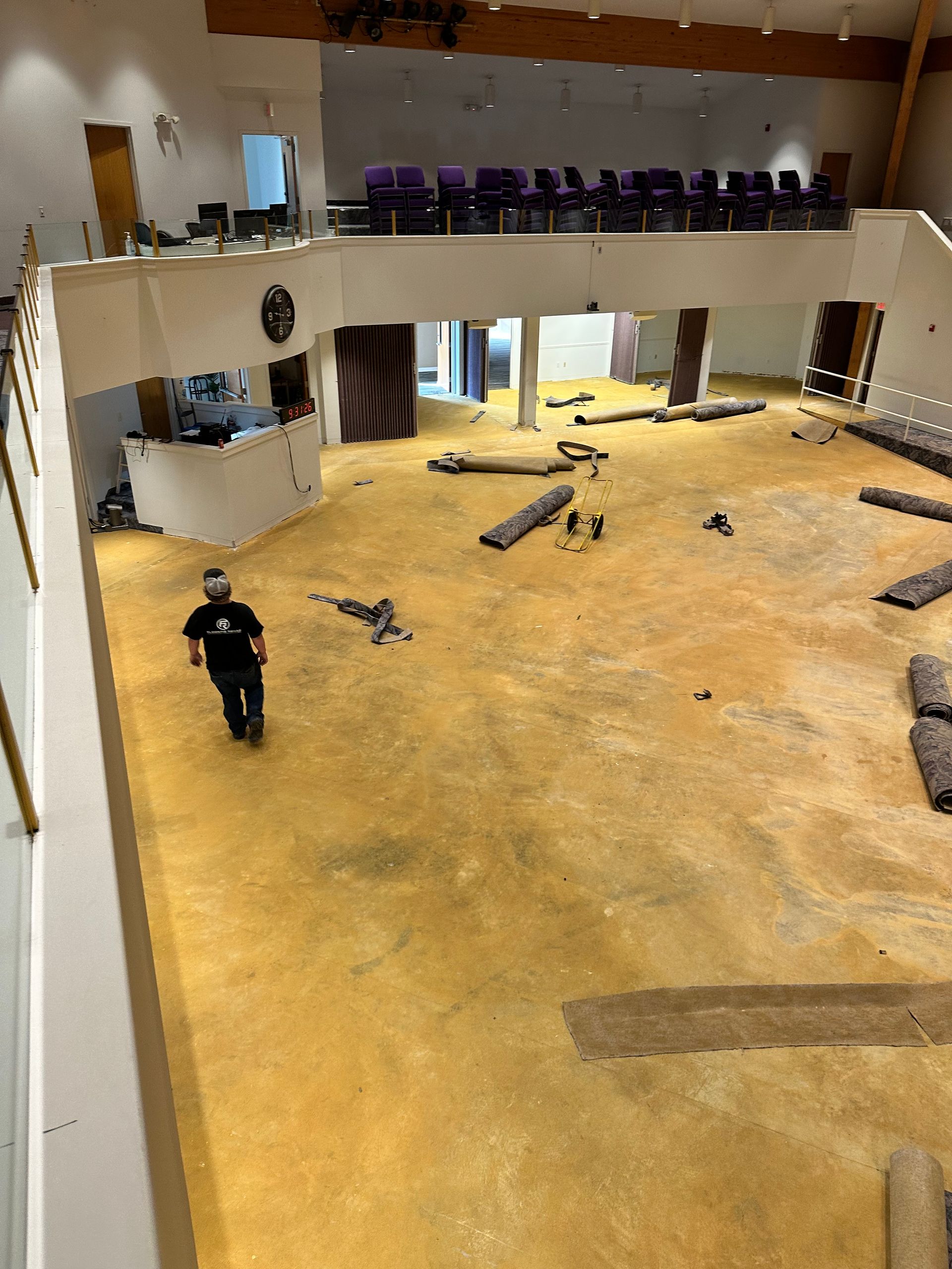 A high-angle shot shows a large, bare room with tan flooring, scattered rolled carpets, and a worker walking across it.