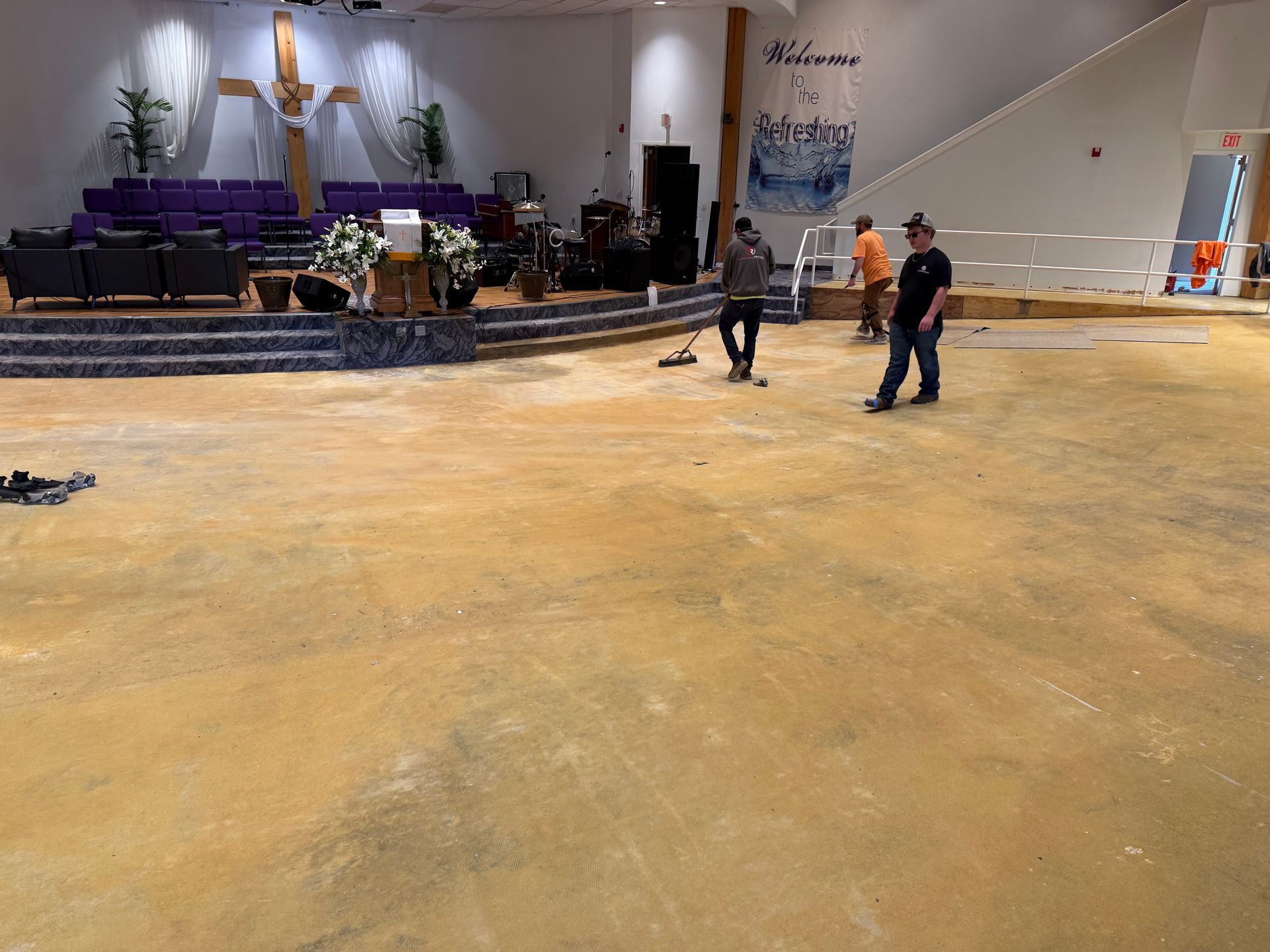 Three workers prepare a large, empty church floor for new carpeting, with the altar and purple seating visible in back.