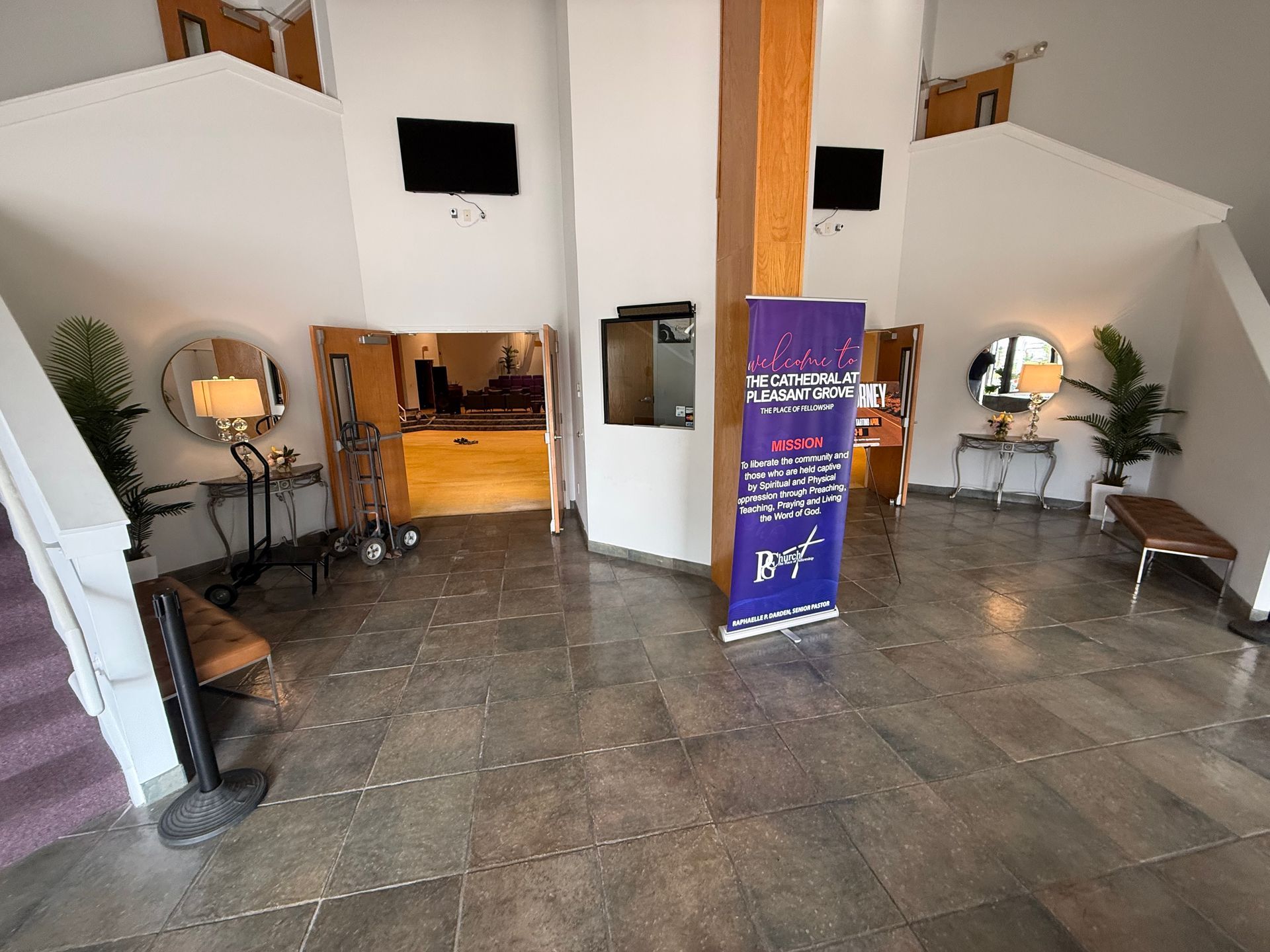 A brightly lit lobby with stone floors, two staircases, decorative mirrors, a wooden pillar, and a purple display banner.