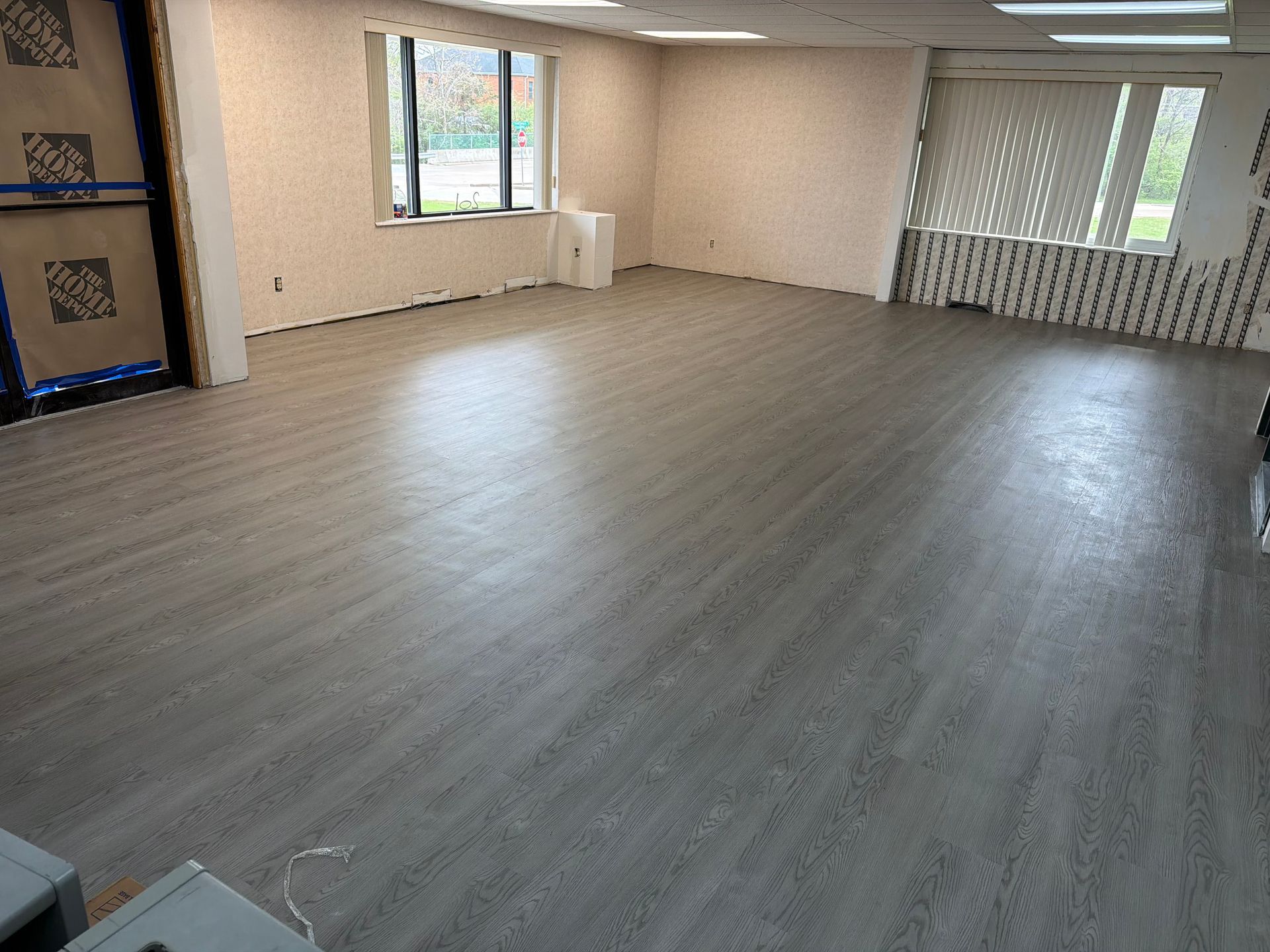 A spacious, empty room featuring light wood-look vinyl flooring, beige walls, and large windows with vertical blinds.