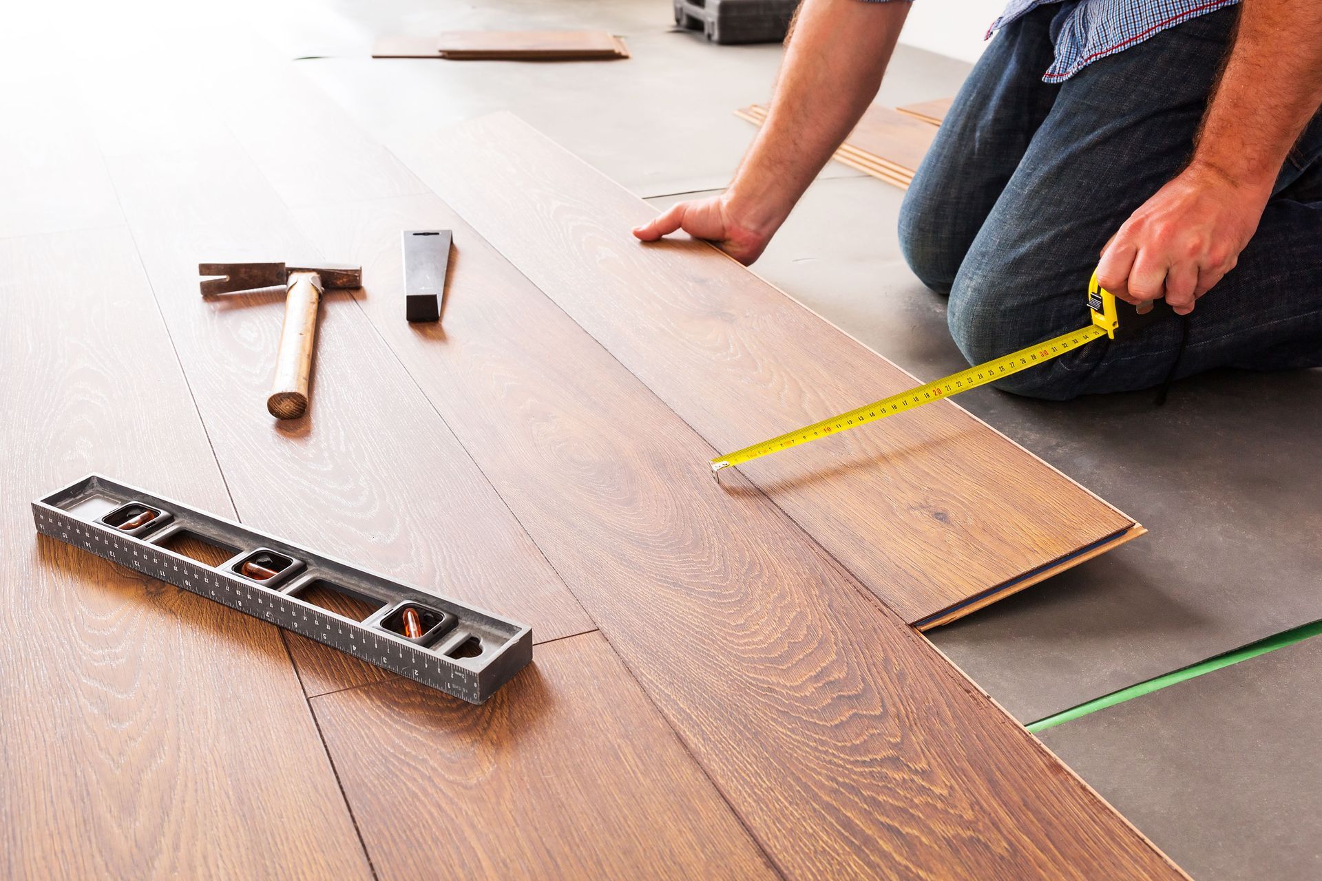 A person installing wood laminate flooring, using a measuring tape, spirit level, and hammer nearby.