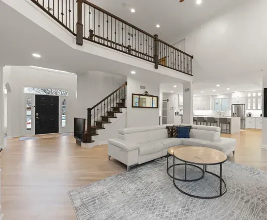 An open-concept living area with a white sofa, round wooden coffee table, staircase with black railings, and a kitchen.