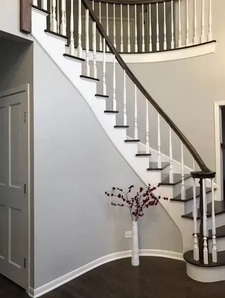 A curved wooden staircase with white spindles and dark steps, featuring a tall vase with red floral branches underneath.