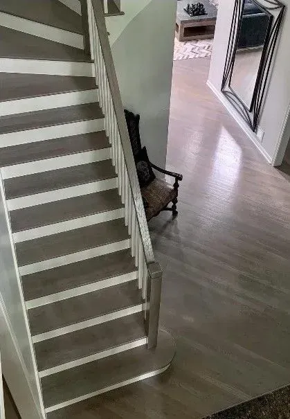 A high-angle view of a staircase with grey treads and white risers, leading to a hallway with wood floors and a mirror.
