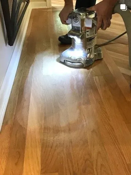 A person uses a floor sander with an integrated light to refinish a hardwood floor along a wall.