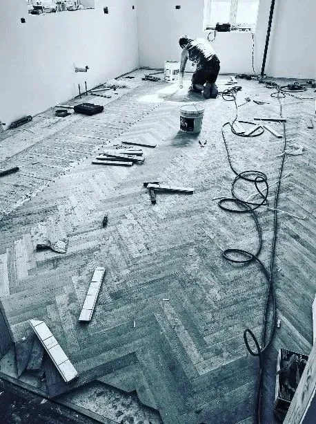 A person kneels while installing a herringbone wood floor in a room under construction, with tools scattered nearby.