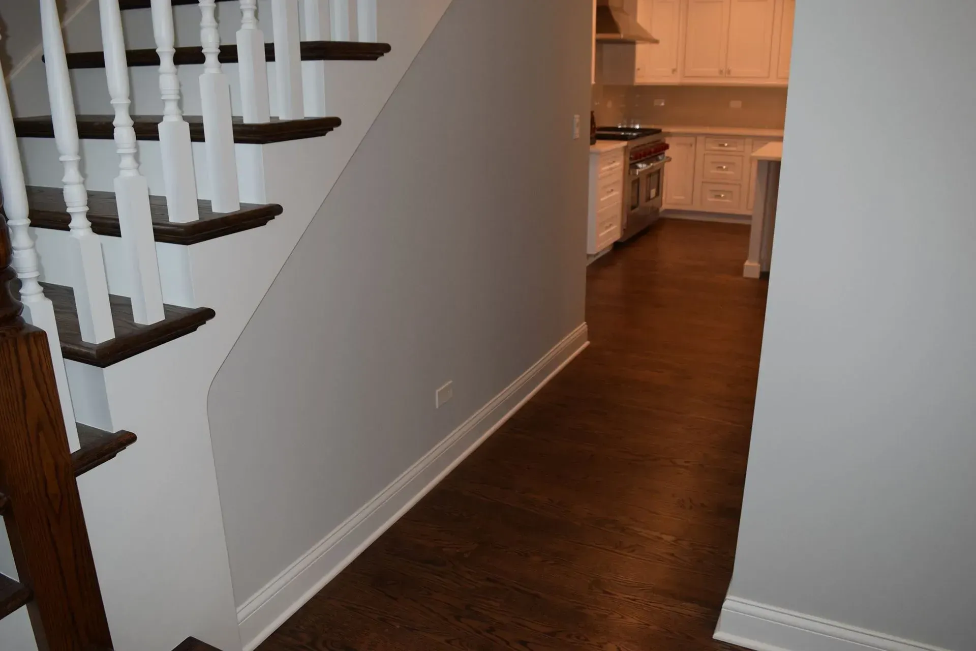 A hallway with a wooden staircase on the left and a view into a bright white kitchen in the background.