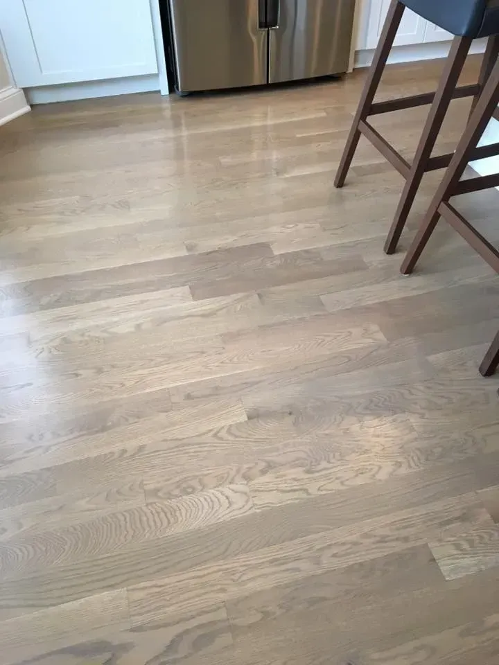 Light-colored hardwood flooring in a kitchen, with a stainless steel appliance and bar stool legs visible in the background.